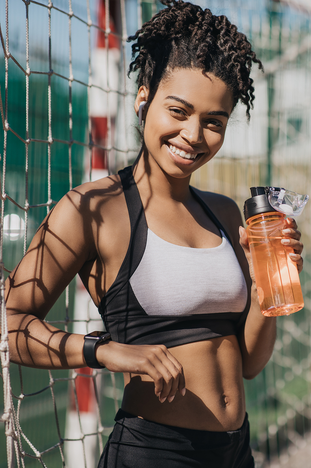 Fitness blogger and sports coach, workout outdoor in city. Smiling millennial african american lady in sportswear with headphones shows modern bottle of water with free space at stadium, vertical